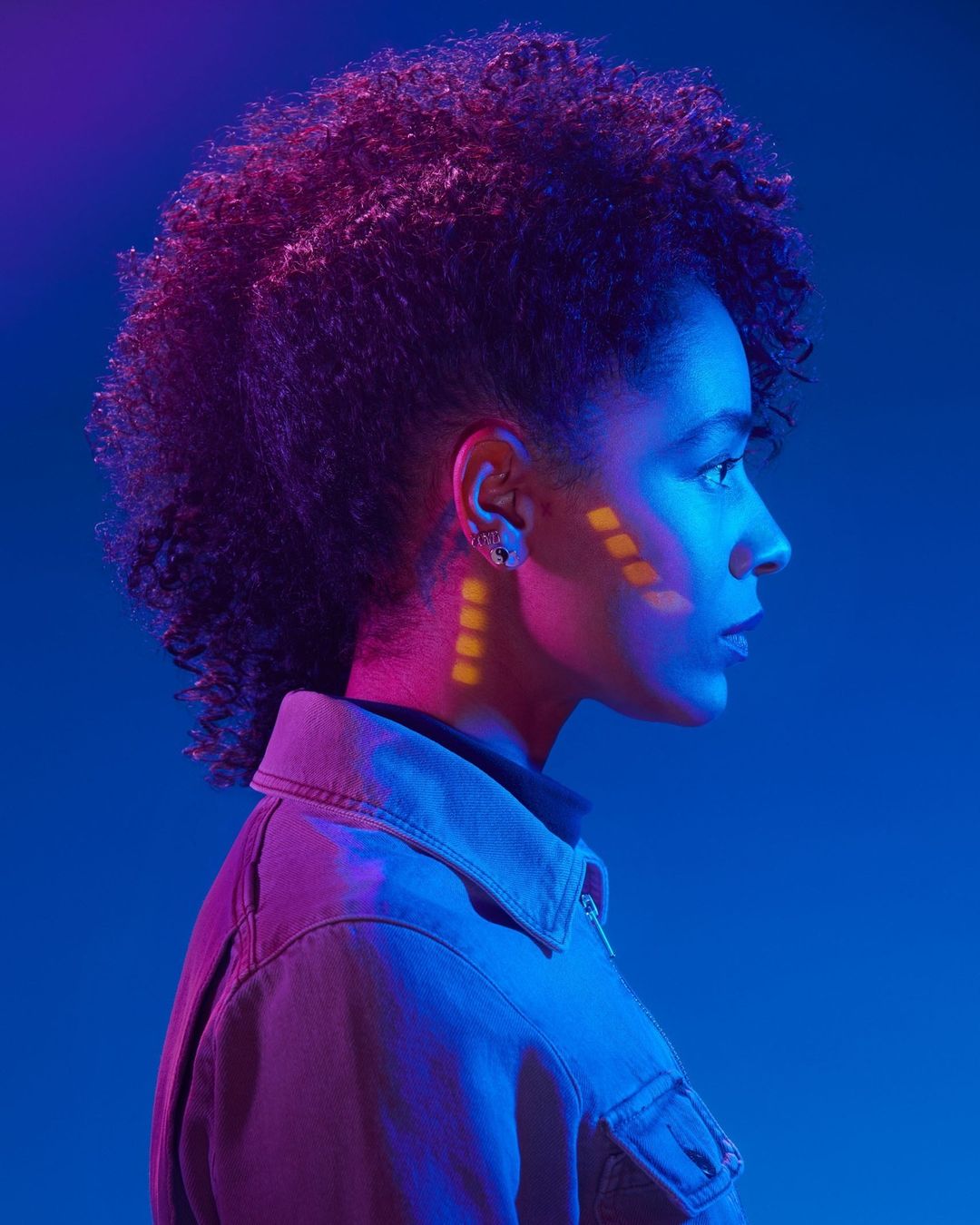 Side profile portrait of a woman with afro hairstyle, geometric makeup, wearing a jacket, illuminated by pink and blue lighting.